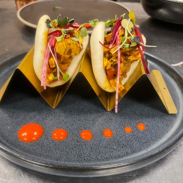 Two Indian bao buns with paneer, microgreens, and pickled onions, served on a dark plate with orange sauce.