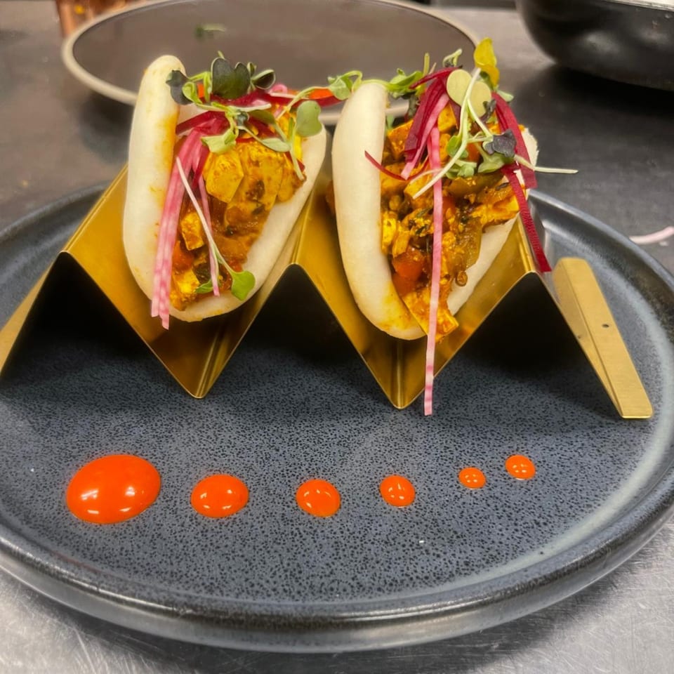 Two Indian bao buns with paneer, microgreens, and pickled onions, served on a dark plate with orange sauce.