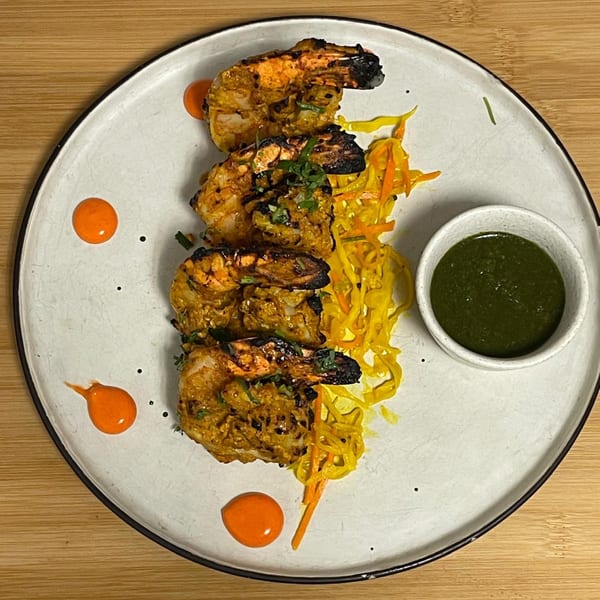 Tandoori shrimp with yellow noodles, orange sauce dots, and green chutney on a white plate.