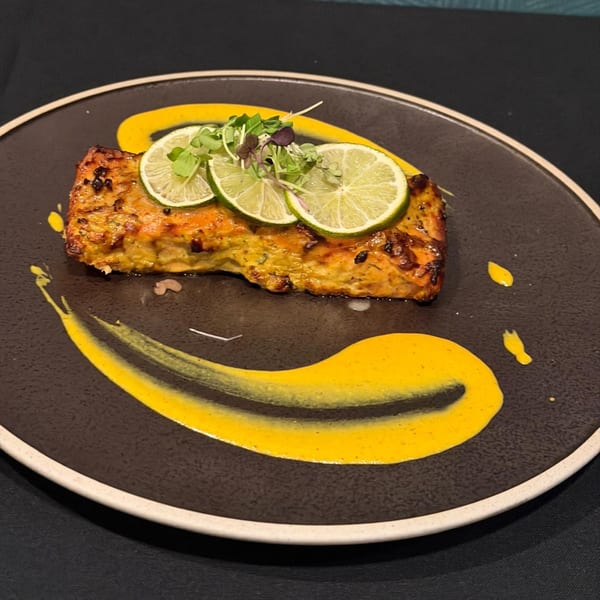 Indian-spiced salmon with lime slices and microgreens, artfully plated with yellow sauce on a dark dish.